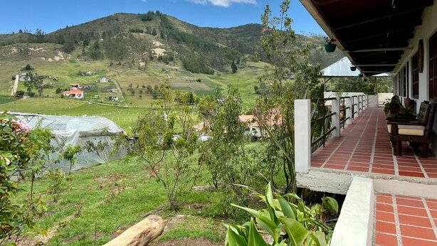 A terrace with a view of a green valley and hills, a covered outdoor seating area, and a greenhouse.