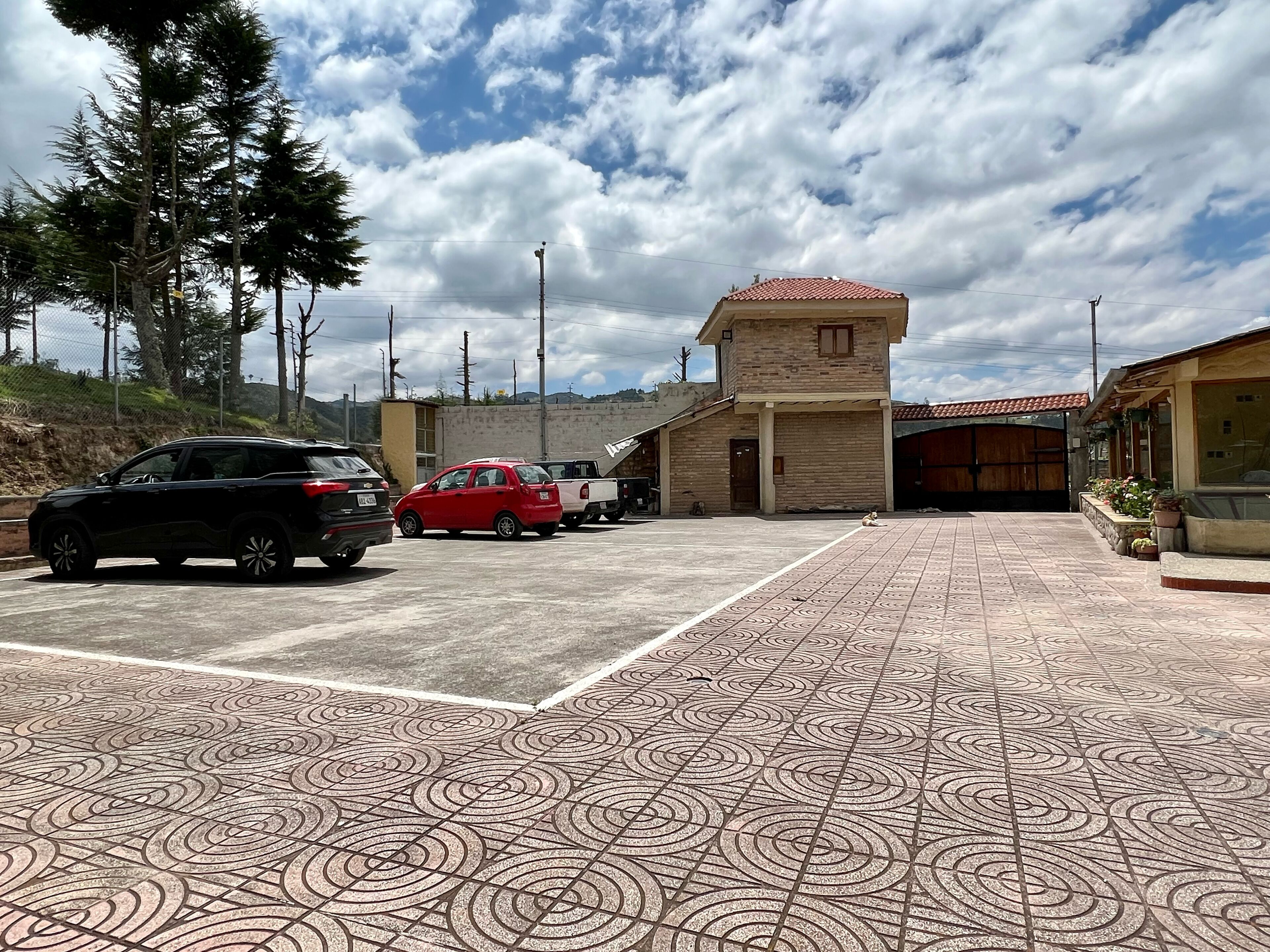 Un estacionamiento con suelo de baldosas estampadas, un edificio de techo en teja roja y algunos coches aparcados.