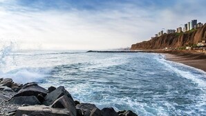 En kustvy med en stenig strand, vågor som bryter mot stranden och en stadsbild i bakgrunden.