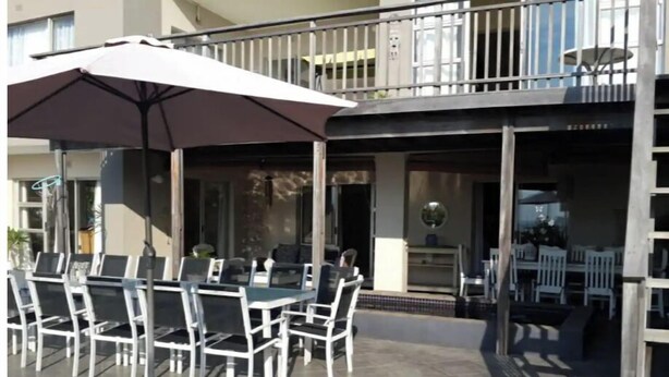 En udendørs terrasse med spisebord og stole, en stor parasol og en balkon med gelænd.