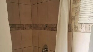 Un petit espace de douche carrelé, avec un rideau blanc, une pomme de douche visible et un lavabo doté d’un bac bleu.