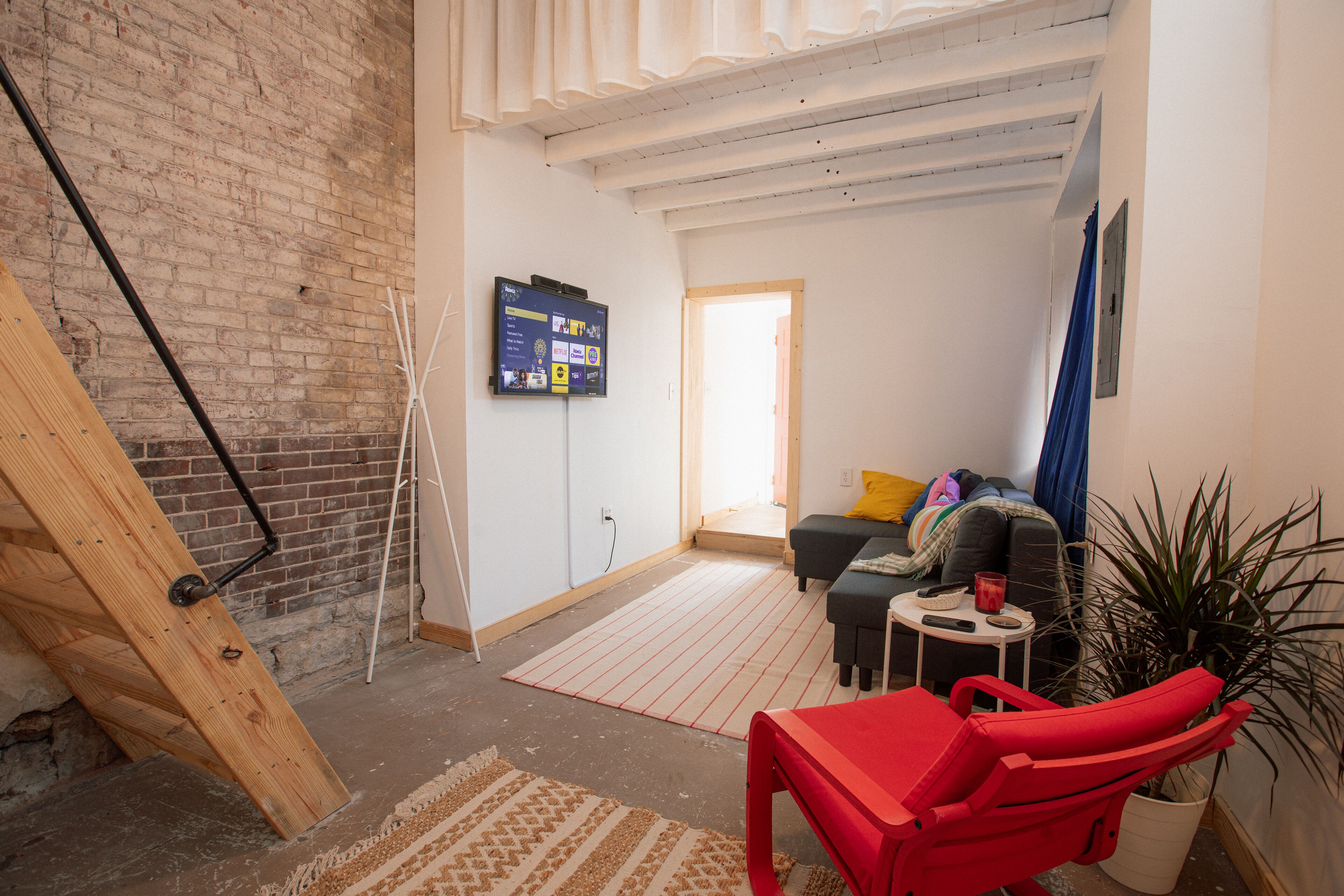 Een kamer met een bakstenen muur, een houten trap, een flatscreen televisie, een bank, een rode stoel en een potplant.