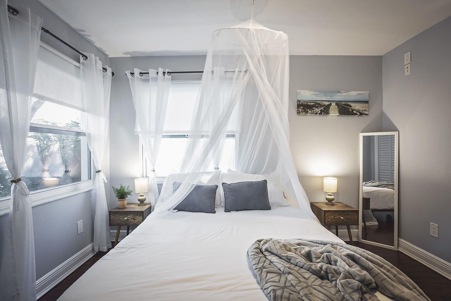 A bedroom with a bed, a bedside table, a nightstand, a window with sheer curtains, a bedside lamp, a mirror, and a wall-mounted picture.
