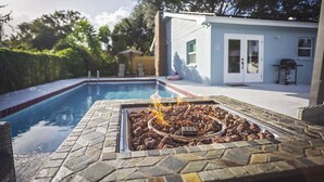 Ein Grillbereich am Pool mit Feuer, umgeben von einer Stein-Terrasse, mit Pool und einem Gebäude im Hintergrund.