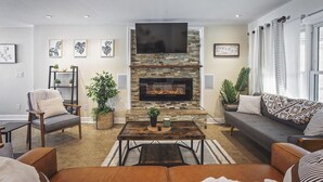 Ein Wohnzimmer mit Kamin aus Stein, einem flachbildfernseher darüber, einer Couch, einem Couchtisch und einem Bücherregal.