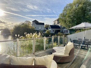 A deck with outdoor furniture, a view of a river, and houses in the background. - Large 4 Bedroom Lakeside Holiday lodge in the Cotswold. Sleeps 8-10 (South Cerney)