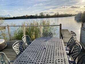 A metal table with chairs on a wooden deck overlooking a calm lake, with a wooden railing and vegetation in the background. - Large 4 Bedroom Lakeside Holiday lodge in the Cotswold. Sleeps 8-10 (South Cerney)