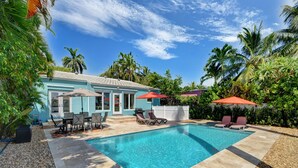 A swimming pool with lounge chairs and umbrellas, surrounded by palm trees and a house with a light blue exterior.