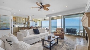 A spacious living area with a large window overlooking the ocean, a ceiling fan, and a kitchen in the background. - Luxury Oceanfront Villa w/ Resort Style Amenities (Oceanside)