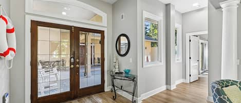A modern interior with a wooden floor, a glass door entrance, a round mirror, and a small table with decorative items.