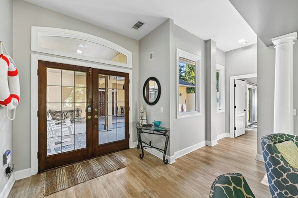 A modern interior with a wooden floor, a glass door entrance, a round mirror, and a small table with decorative items. - Beautiful Home w/ Pool & Game Room (Hilton Head Island)