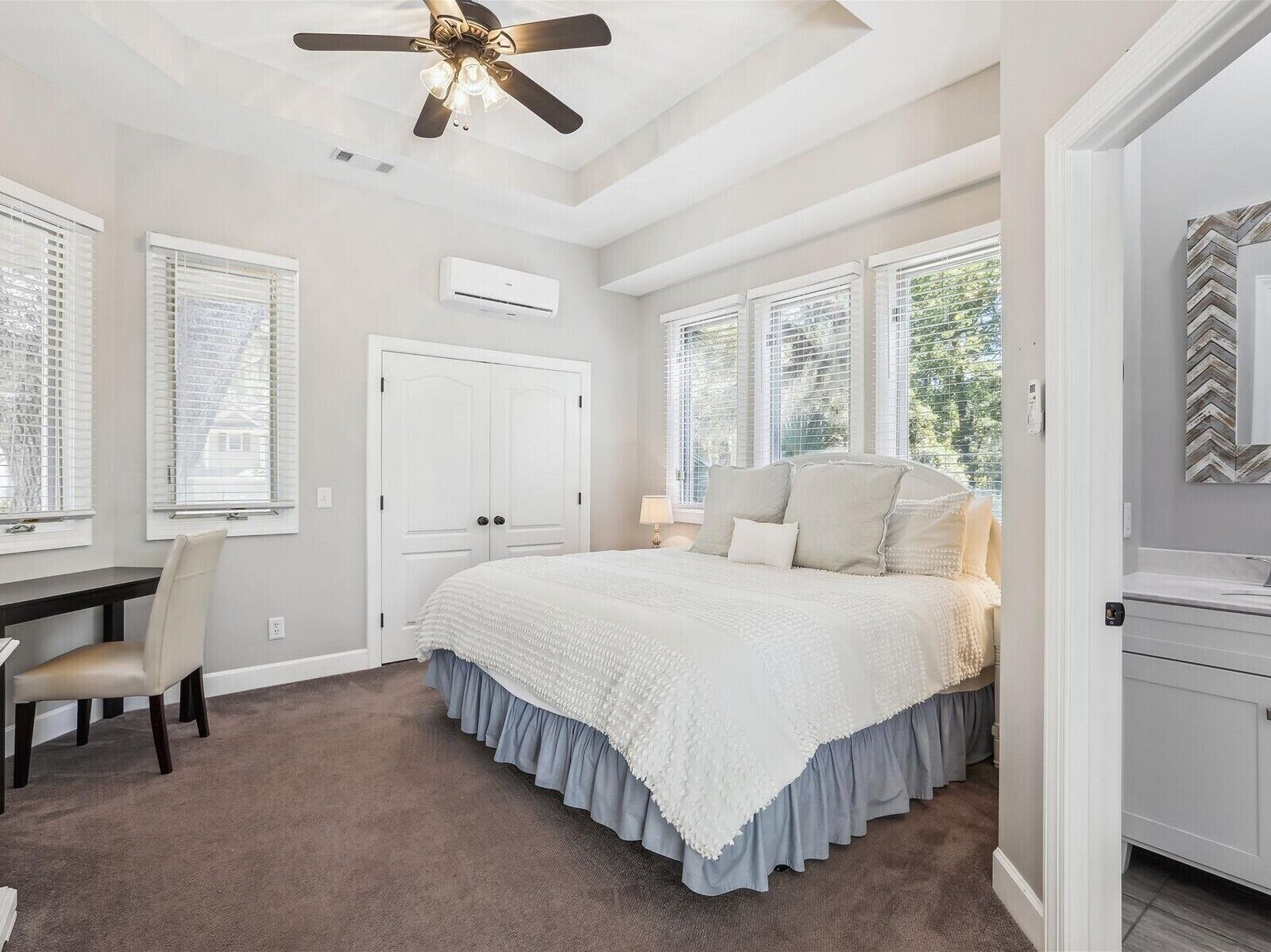 A bedroom with a bed, a desk, a chair, a ceiling fan, and large windows.