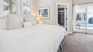 Een slaapkamer met een groot bed, een nachtkastje, een lijst met een afbeelding, en uitzicht op een zwembad door een open deur.