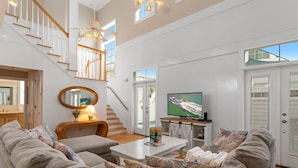 A spacious living room with a high ceiling, a staircase, a television, and a large window.