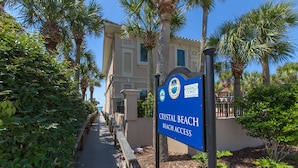 A sign for Crystal Beach Beach Access with a pathway leading to a building surrounded by palm trees.