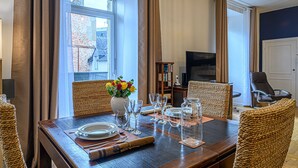 Un coin repas avec une table en bois dressée pour quatre personnes, entourée de chaises en osier. Un vase rempli de fleurs est posé sur la table, et une bibliothèque se trouve à l’arrière-plan.