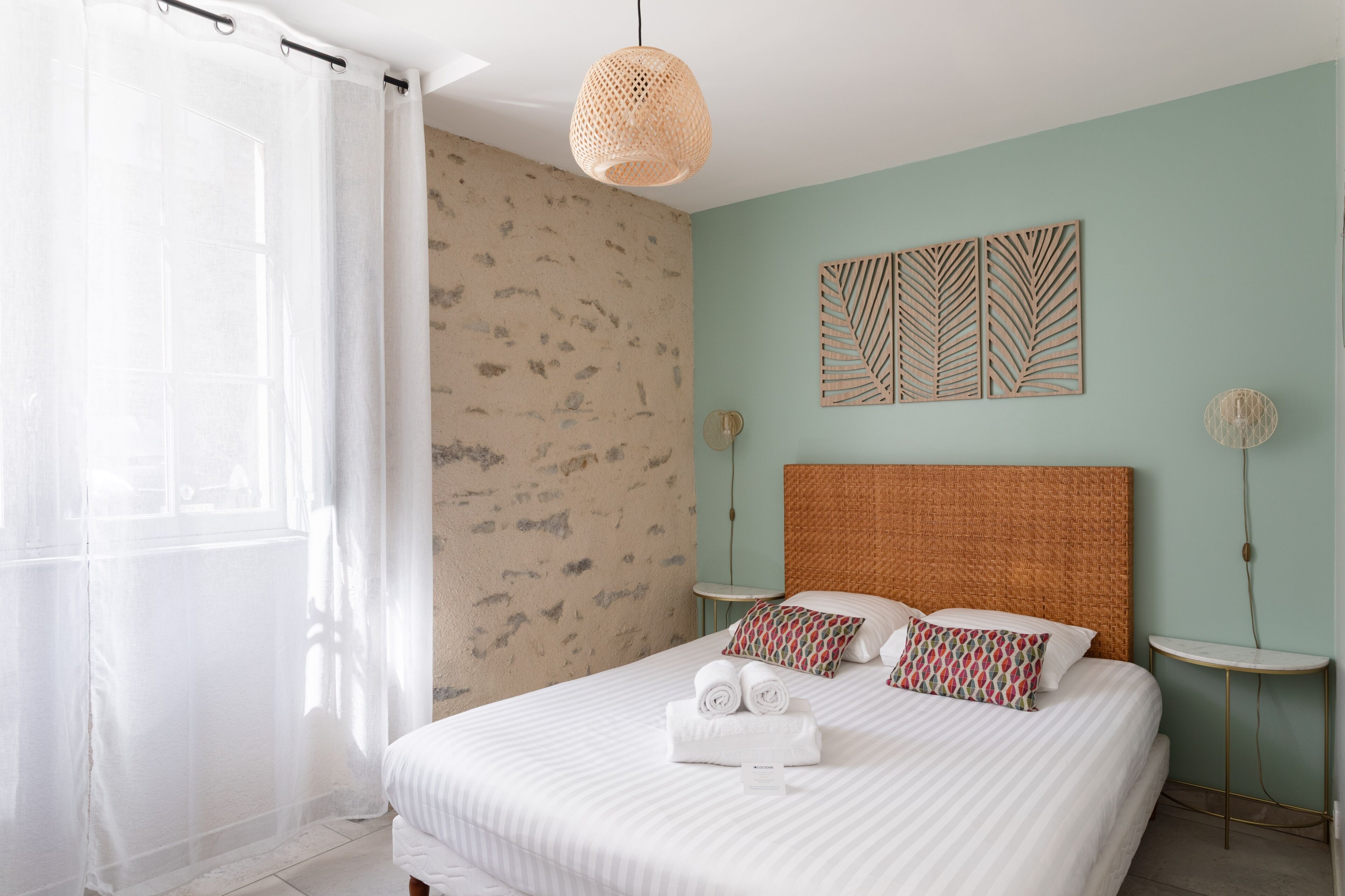 Een slaapkamer met een bed, een hoofdbord, een wand met een bladmotief, een nachtkastje en een hanglamp.