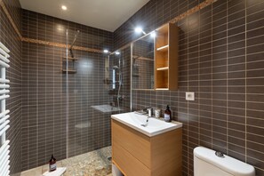 A modern bathroom with dark tiled walls, a wooden vanity, a white sink, a glass shower enclosure, and a mirror above the vanity.