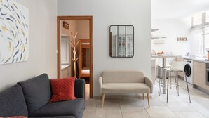Ein modernes Wohnzimmer mit einer Couch, einer Bank, einer Küchenzeile mit Waschmaschine und einem Spiegel an der Wand.