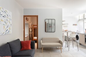 A modern living room with a sofa, a bench, a kitchen area with a washing machine, and a mirror on the wall.