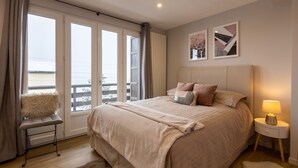 A bedroom with a large bed, a nightstand, a fluffy chair, and a view of the sea through large windows. - Orpin - superbe appartement rénové 2 personnes avec grande terrasse plein sud, belle vue (Font-Romeu)