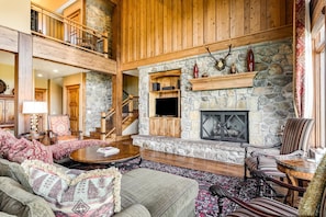 A spacious living room with a stone fireplace, wooden furniture, and a high ceiling with a balcony.