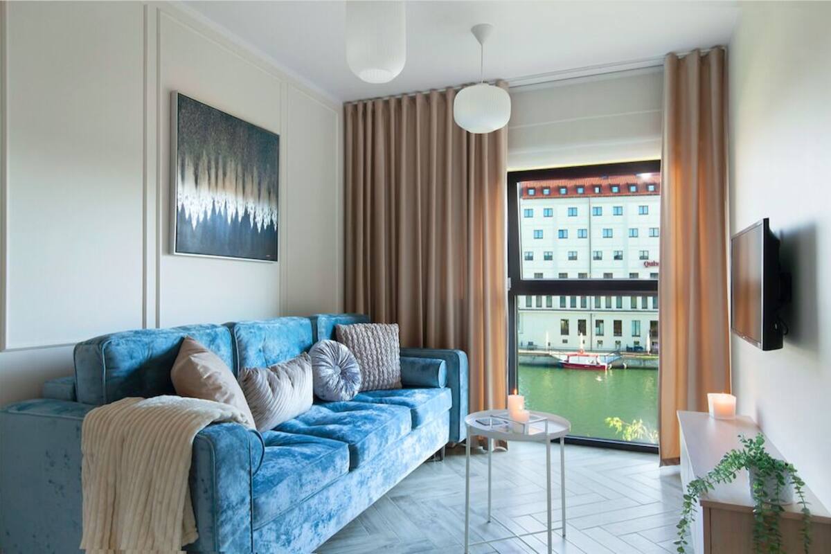 A modern living room with a blue sofa, a flat-screen TV mounted on the wall, and a view of a building across a body of water.