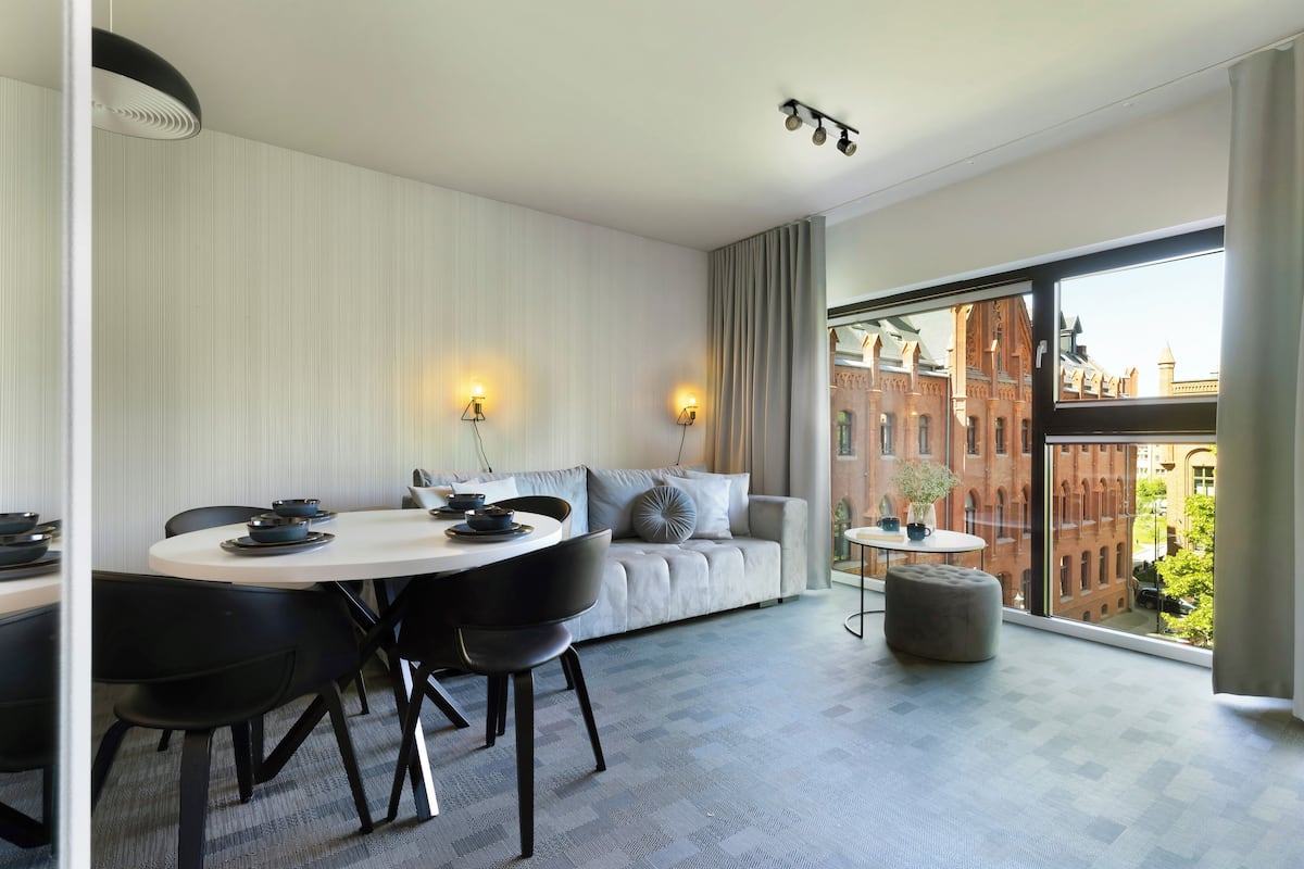 A modern hotel room with a dining area, a sofa, and a large window offering a city view.