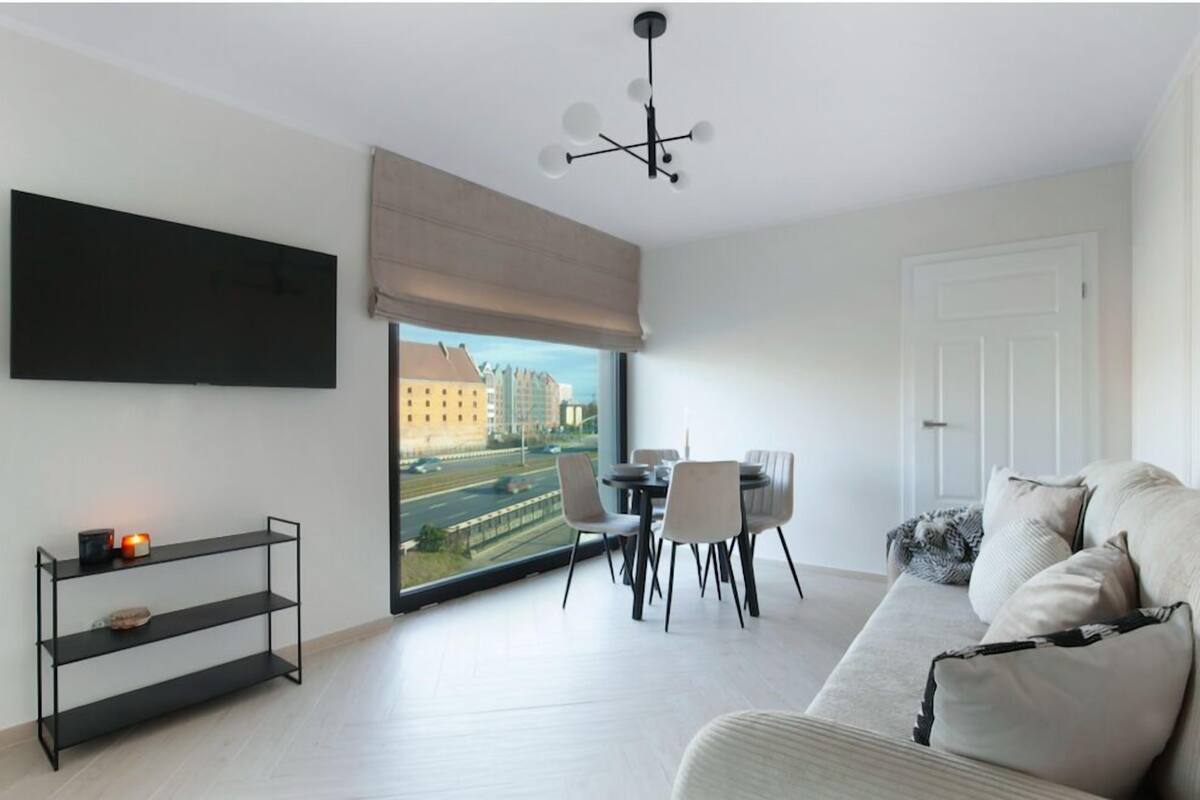 A modern living room with a large window, a flat-screen TV, a dining table with chairs, and a sofa.