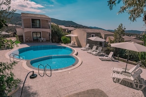A swimming pool with lounge chairs and umbrellas, surrounded by buildings with a mountainous backdrop. - Flat with Pool in Badus Resort (Badesi)