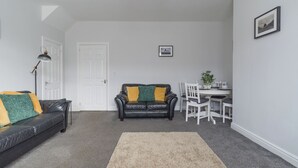 A living room with a black leather sofa, a beige rug, a white door, and a dining area with a table and chairs. - Hawthorn- Great 2 bed home in Ashington, Northumberland (Ashington)
