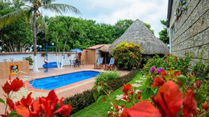 A swimming pool with a thatched-roof structure, surrounded by lush greenery and a well-maintained garden. - Portoazul Casa de Playa (Puerto Colombia)