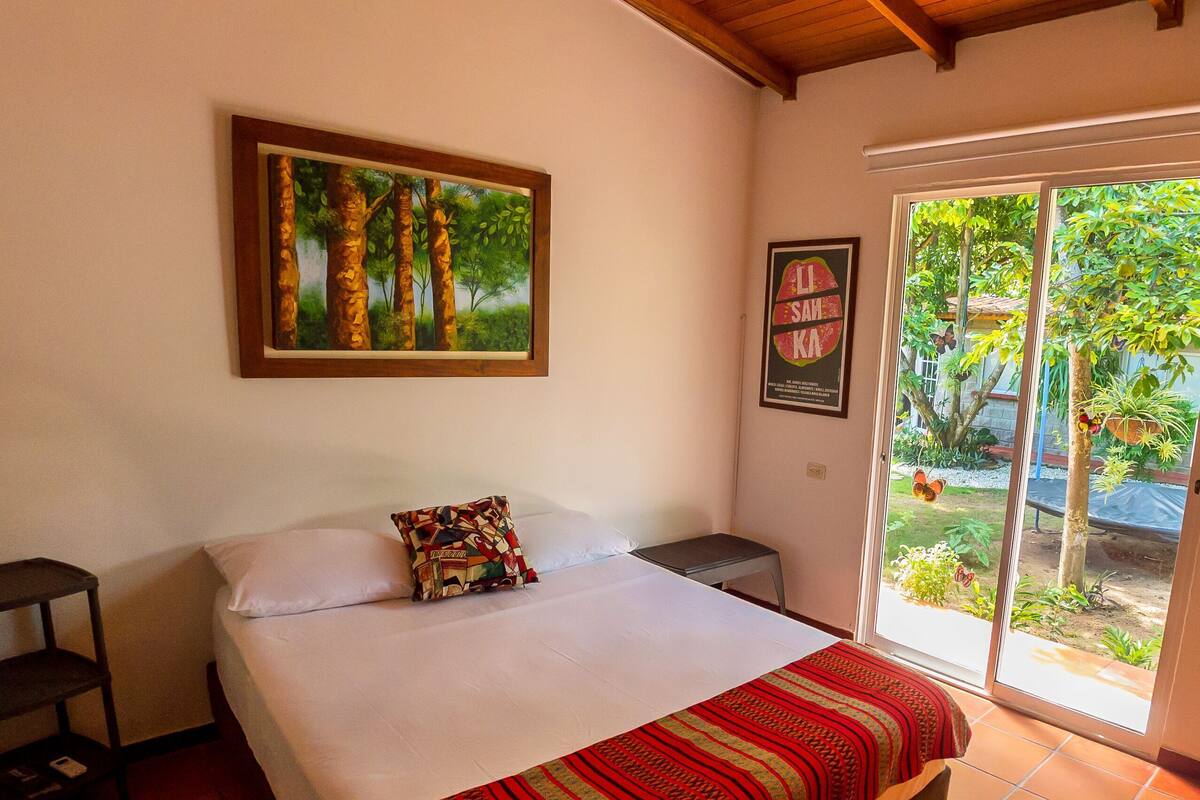 Ein Schlafzimmer mit einem Bett, einem Deckenventilator, einem Fenster mit Blick ins Grüne und einem gerahmten Bild an der Wand.