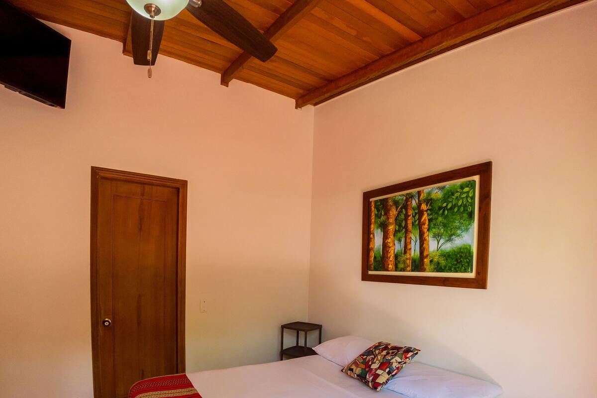 Ein Schlafzimmer mit einem Bett, Deckenventilator, Tür, einem kleinen Tisch und einem Gemälde an der Wand.