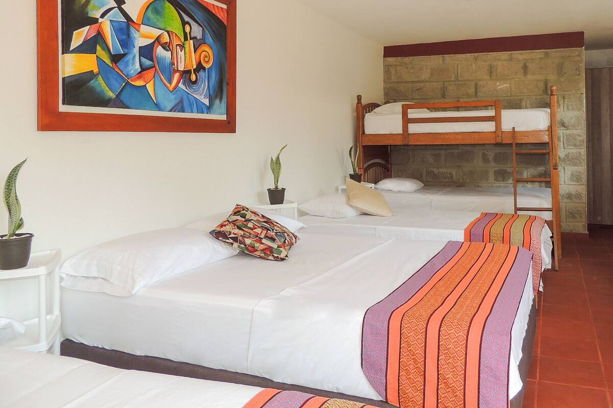 Ein Hotelzimmer mit einem Etagenbett, einem Einzelbett, einem farbenfrohen abstrakten Gemälde und zwei Zimmerpflanzen.