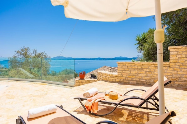 Un espace extérieur lumineux, aménagé pour s’asseoir, avec vue sur la mer, des murs en pierre et une balustrade en verre.