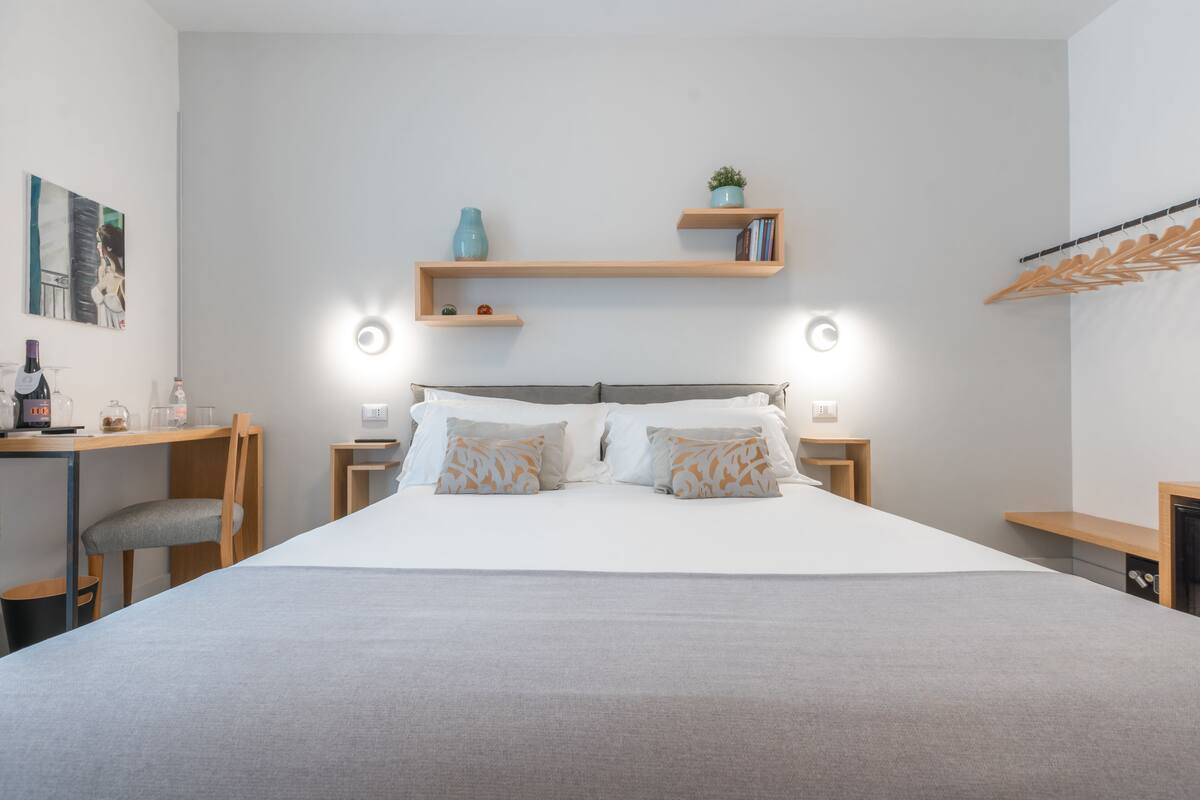 A modern hotel room with a large bed, bedside tables, a desk, and a chair. There are wall-mounted shelves with decorative items and a coat rack.