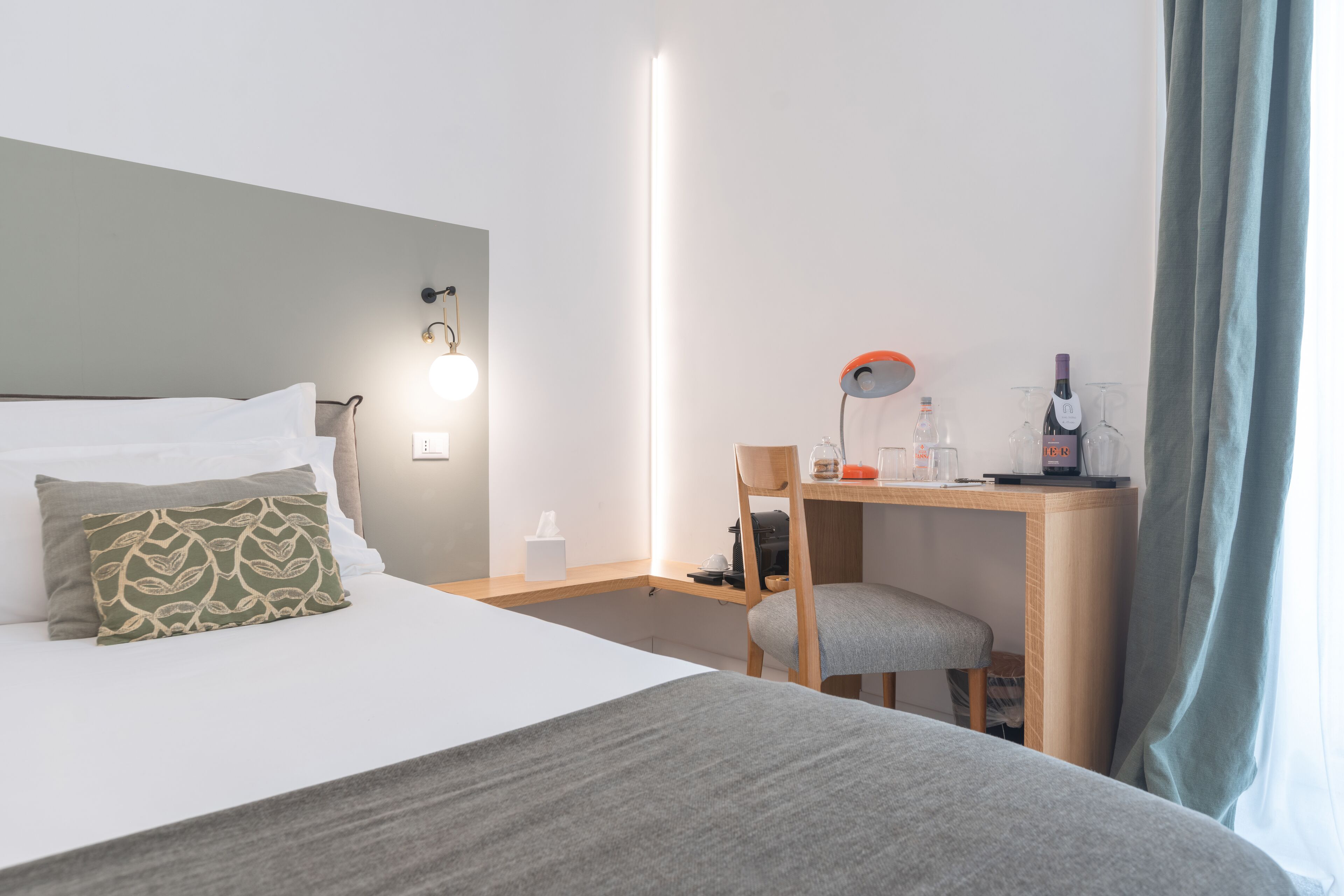 A hotel room with a bed, a desk, a chair, and a nightstand with a lamp and a bottle.