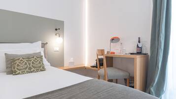 A hotel room with a bed, a desk, a chair, and a nightstand with a lamp and a bottle.