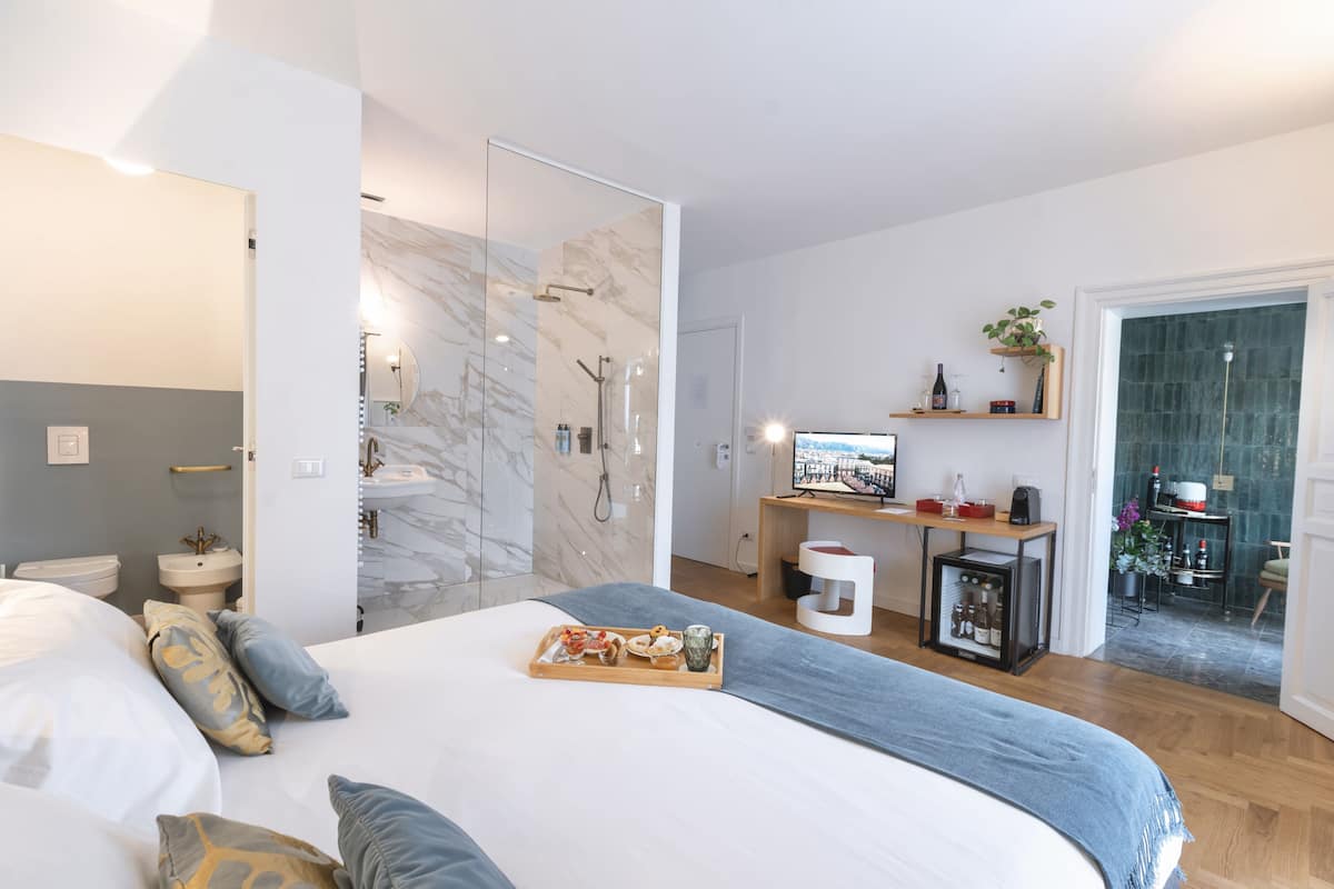A modern hotel room with a large bed, a bathroom with a glass shower enclosure, a small kitchen area, and a dining table with a tray of food.