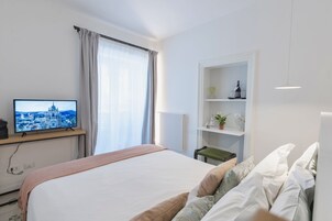Une chambre d’hôtel avec un lit, un téléviseur diffusant une vue sur la ville, une fenêtre avec des rideaux, une étagère avec quelques objets et une chaise.