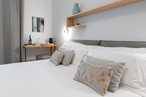 Une chambre d’hôtel bien rangée, avec un lit, des oreillers, un bureau avec une chaise, une étagère avec des objets décoratifs et un tableau au mur.