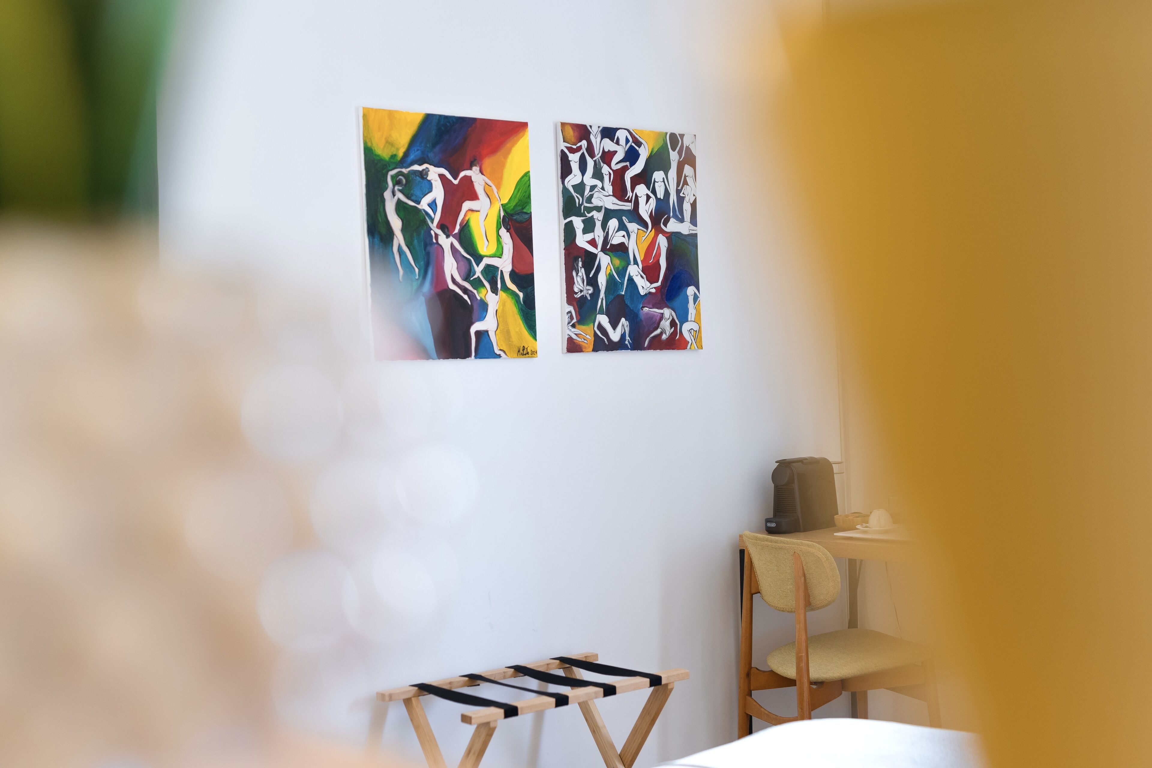 Two abstract paintings on a white wall, a wooden chair, and a small table with a cup.
