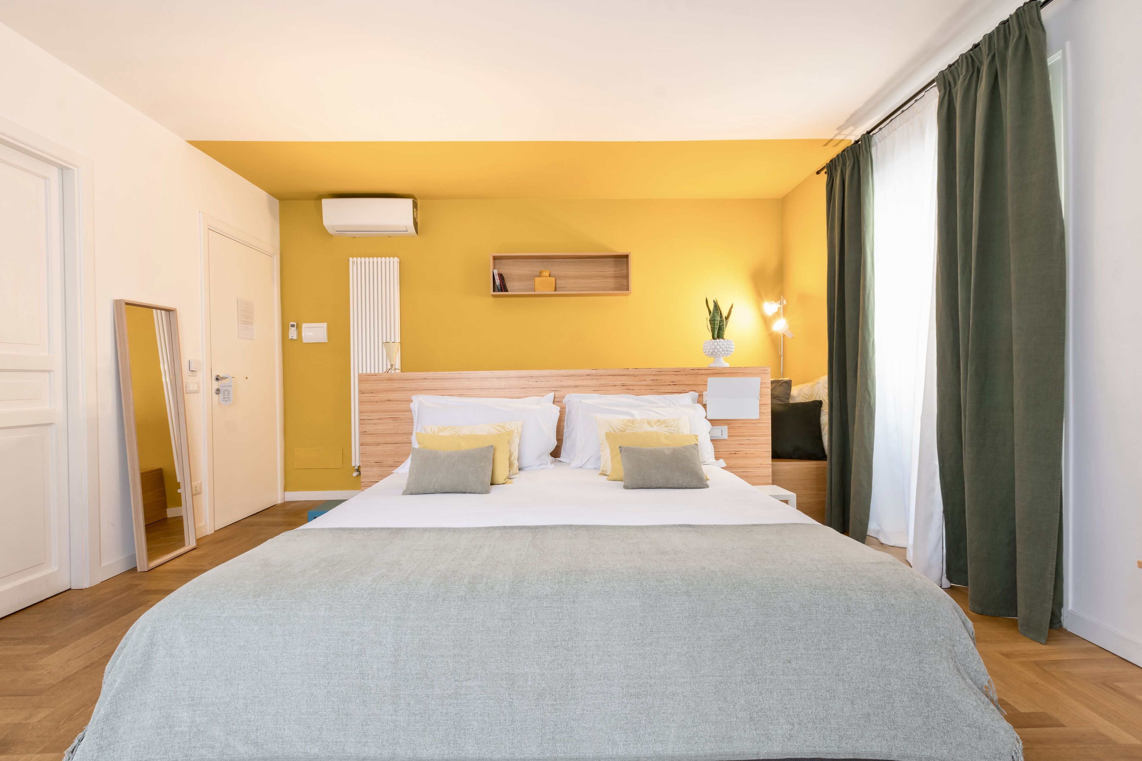 A hotel room with a bed, a bedside table, a lamp, a plant, and a window with curtains.