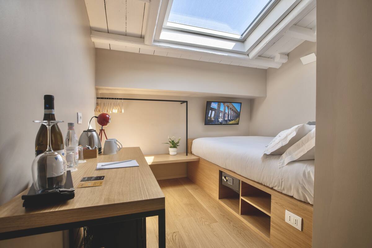 A compact bedroom with a skylight, a bed, a bedside table, a small plant, and a wall-mounted TV.