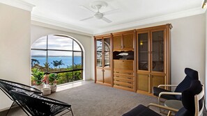 Quarto com uma grande janela em arco com vista para o oceano, um ventilador de teto, um armário de madeira com gavetas e duas cadeiras.