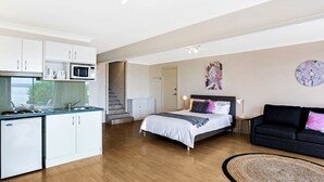 A modern bedroom with a bed, a black sofa, a kitchen area with white cabinets, and a staircase leading to another room. - The Bay- Stunning water views (Nelson Bay)