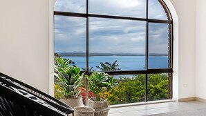 A room with a large arched window overlooking a body of water and distant mountains. There are potted plants in wicker baskets on the windowsill. - The Bay- Stunning water views (Nelson Bay)