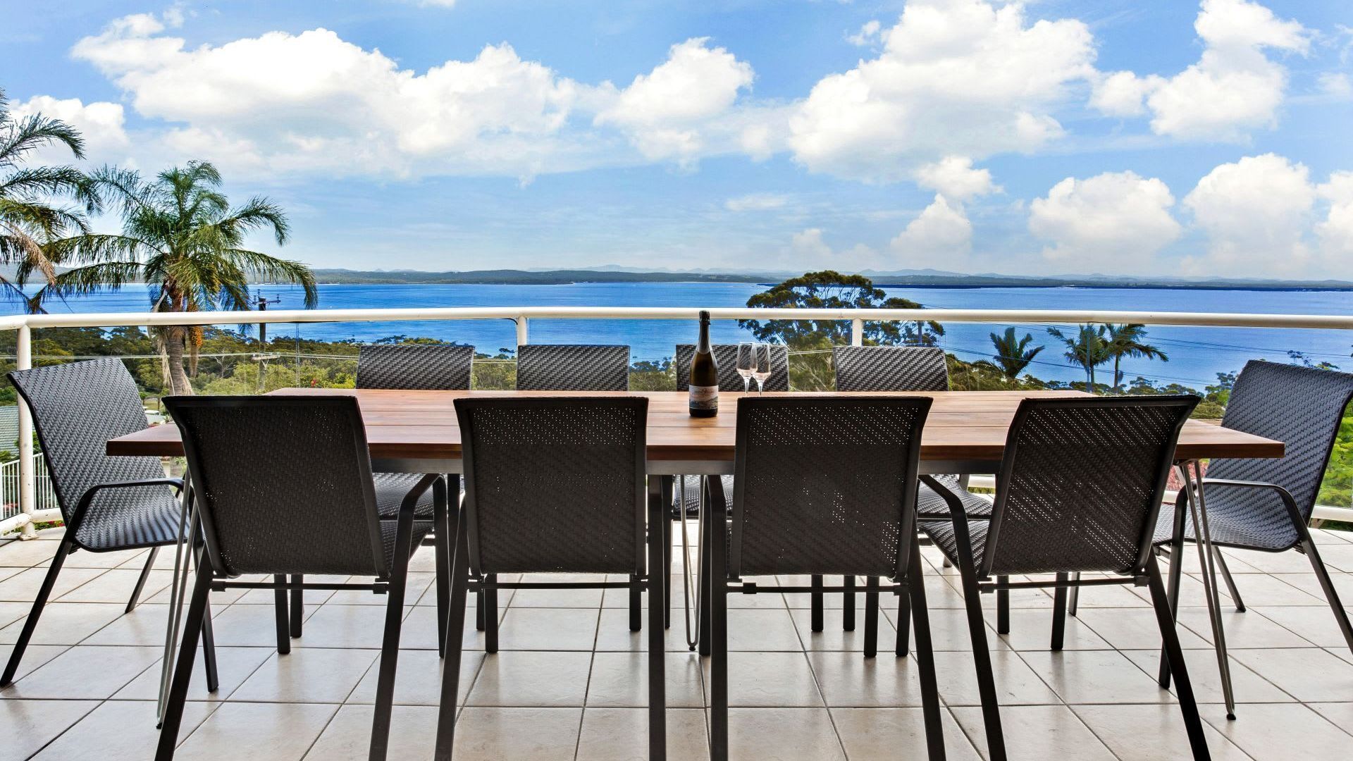 A dining area with a table and chairs, a bottle of wine, and a view of the ocean and mountains.