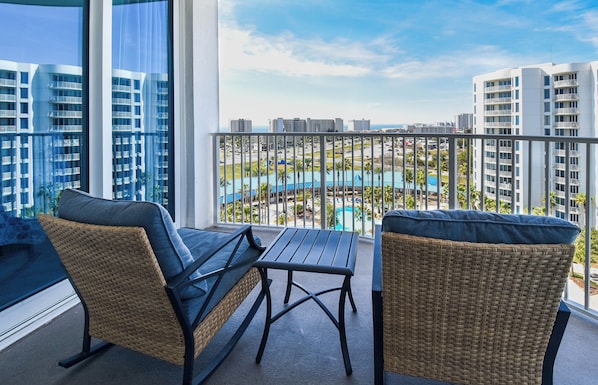 Un balcón con sillas de mimbre y una mesa, con vista a una piscina y edificios.